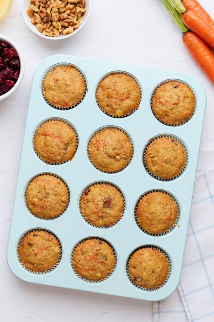 A blue cupcake tray with baked cupcakes in it and carrots in the background.