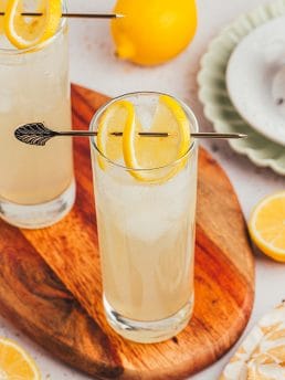 An image of two tall glasses with lemon elderflower mocktail s garnished with lemon slices on a wooden board.