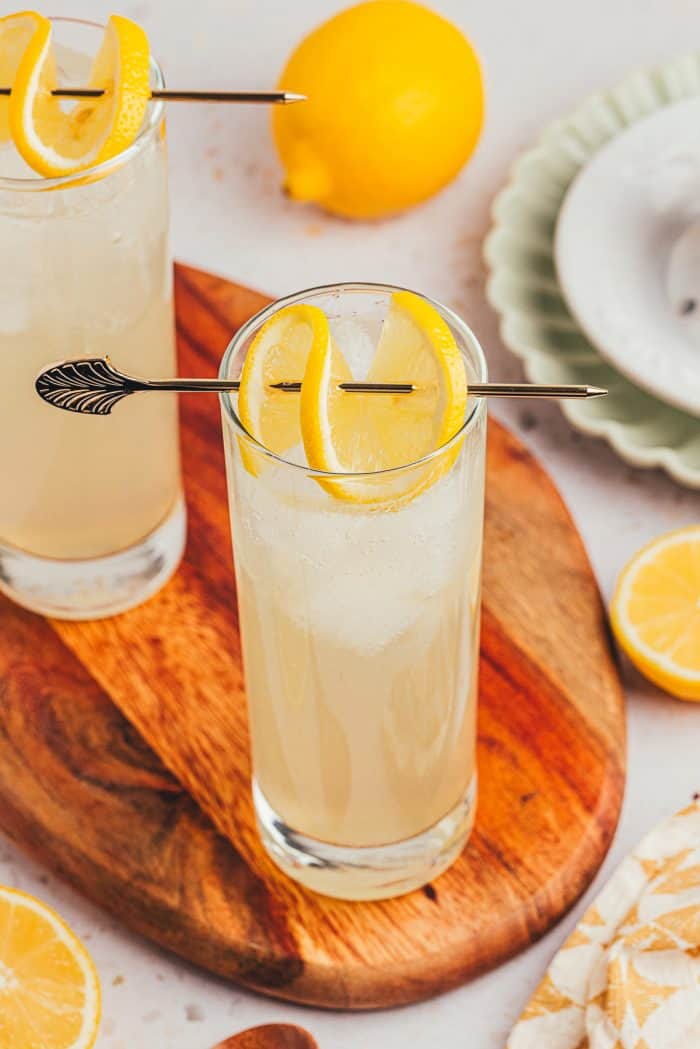 An image of two tall glasses with lemon elderflower mocktail s garnished with lemon slices on a wooden board.