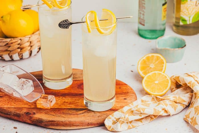 An image of two tall glasses with lemon elderflower mocktail s garnished with lemon slices on a wooden board.