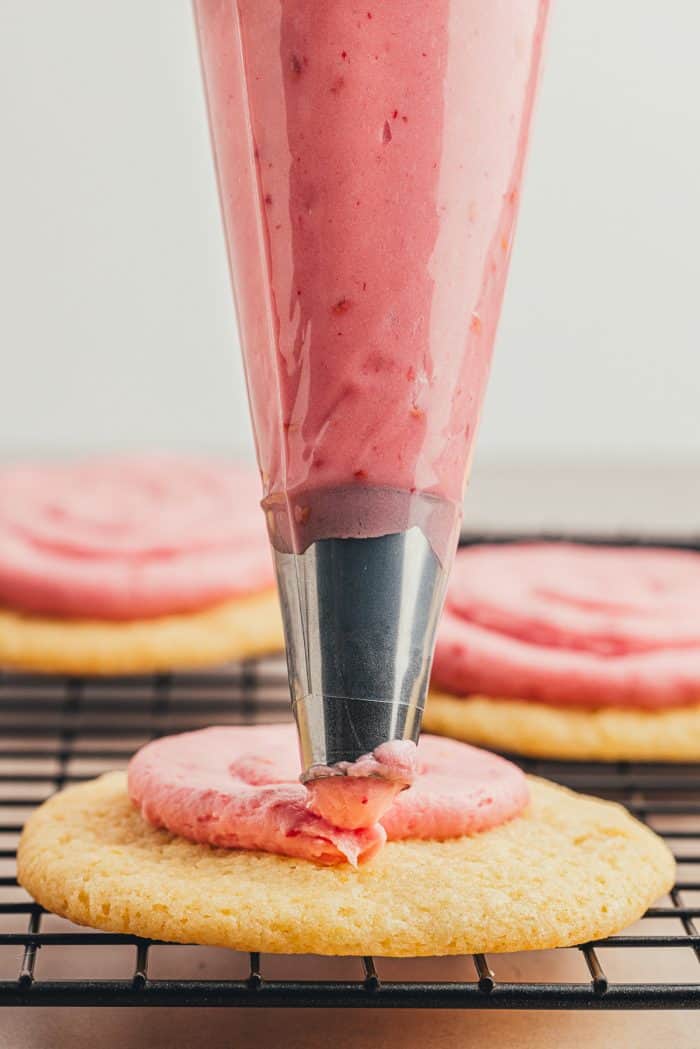 A piping bag, piping pink frosting onto a sugar cookies. 