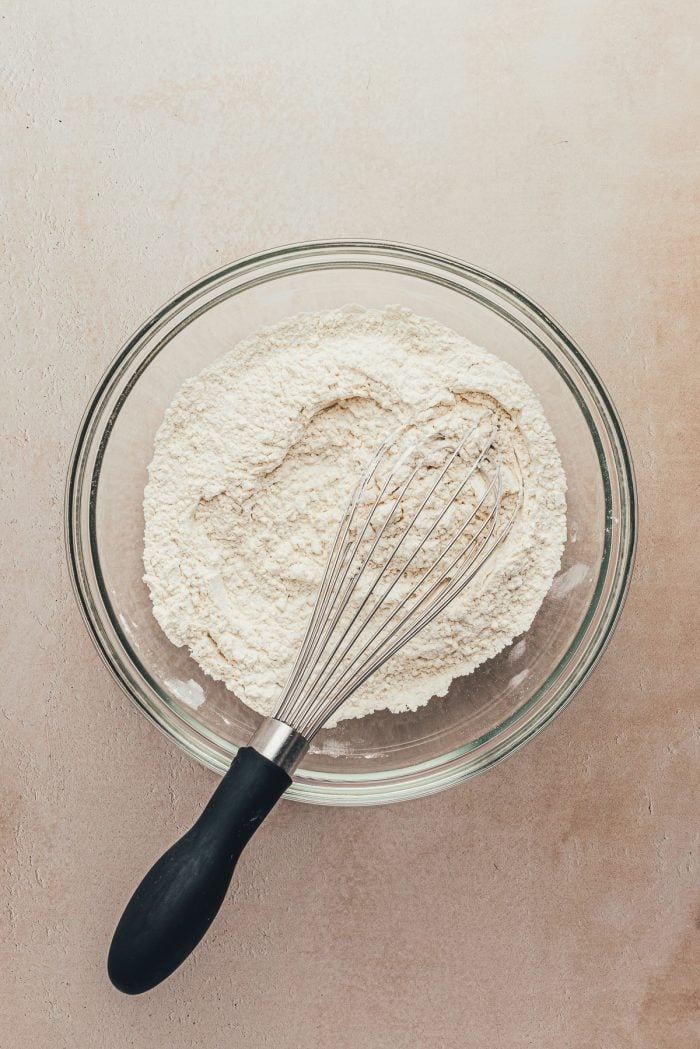 A glass bowl containing flour and a whisk.