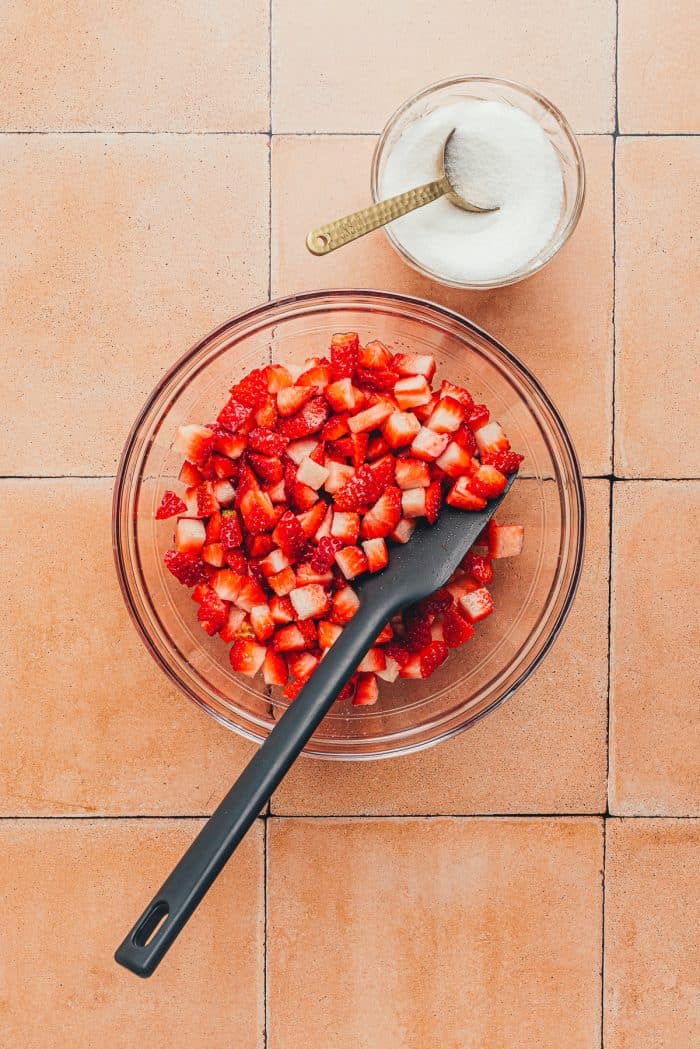 A glass bowl with diced strawberries and a black spatula. 