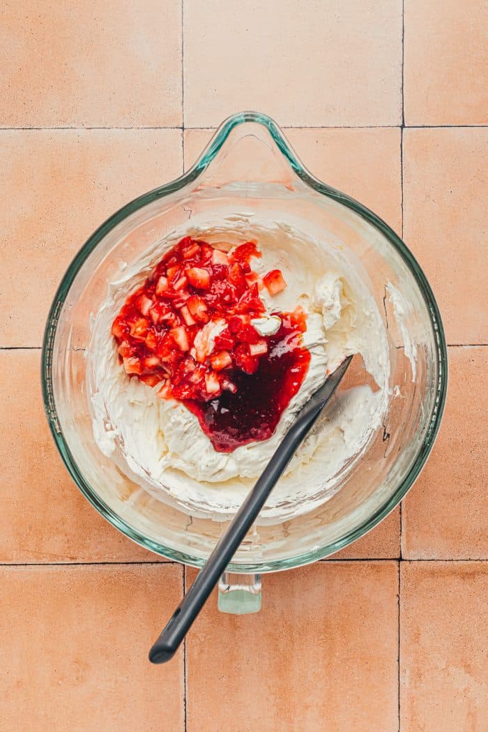 Diced strawberries over the whipped heavy cream mixture with a black spatula in a glass bowl.
