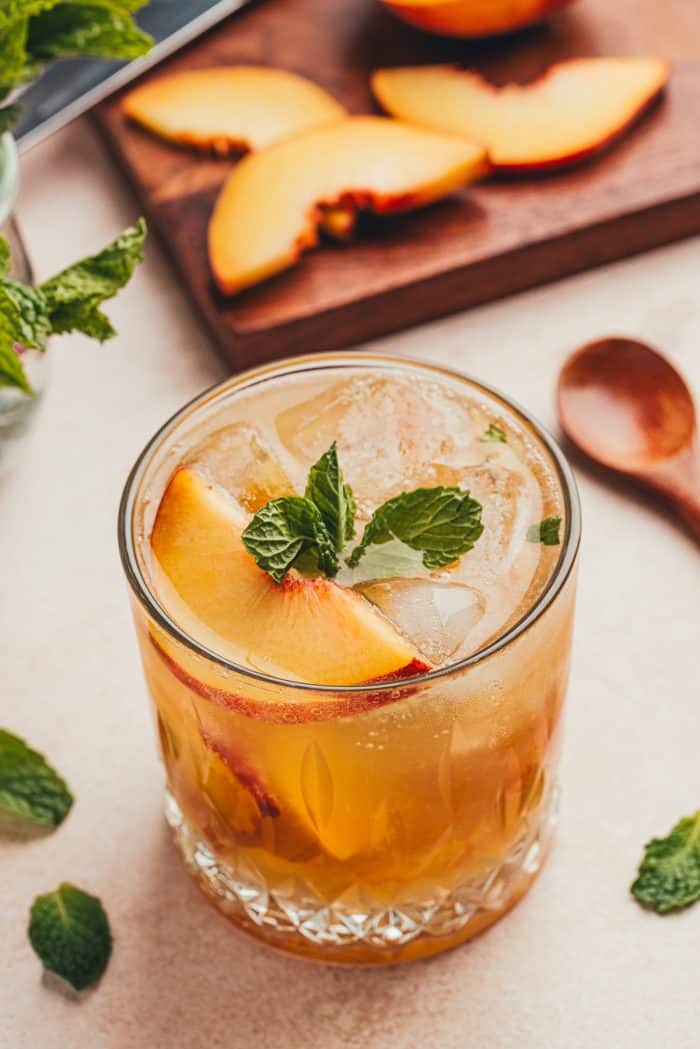 A glass filled with a bourbon peach smash and a wooden board with sliced peaches in the background.