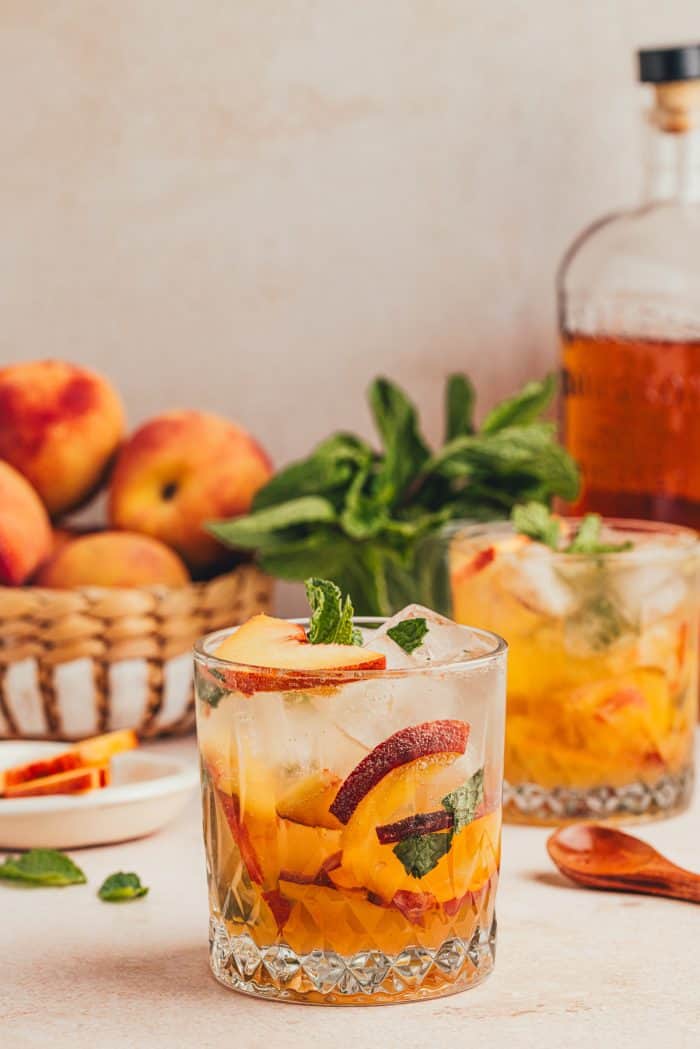 A glass filled with a bourbon peach smash with a bowl of peaches in the background and a bottle of bourbon.