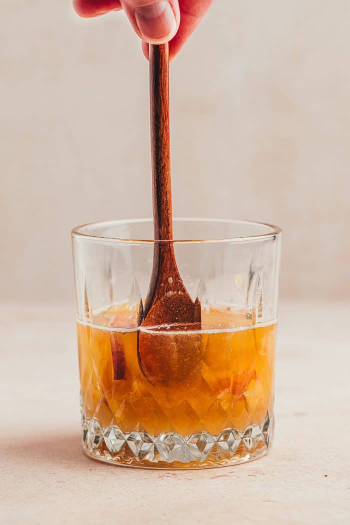 A glass with a wooden spoon and the ingredients for a peach bourbon smash.