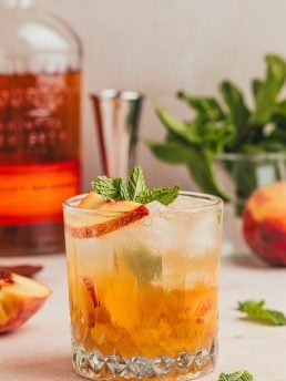 A bourbon peach smash with garnishes of sliced peaches and herbs.