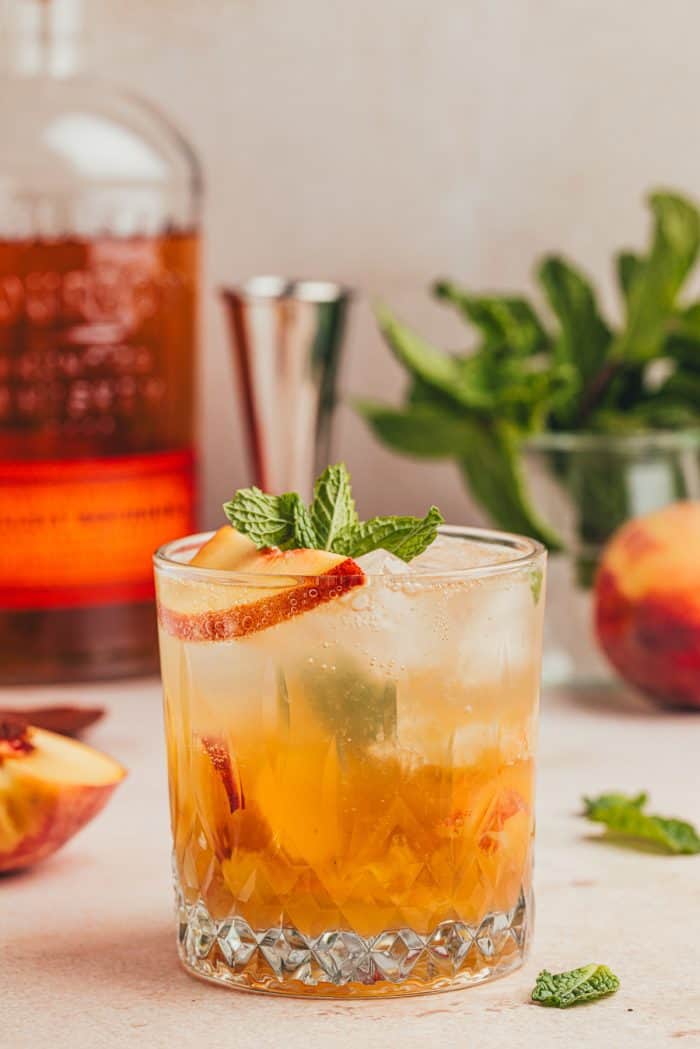 A bourbon peach smash with garnishes of sliced peaches and herbs.