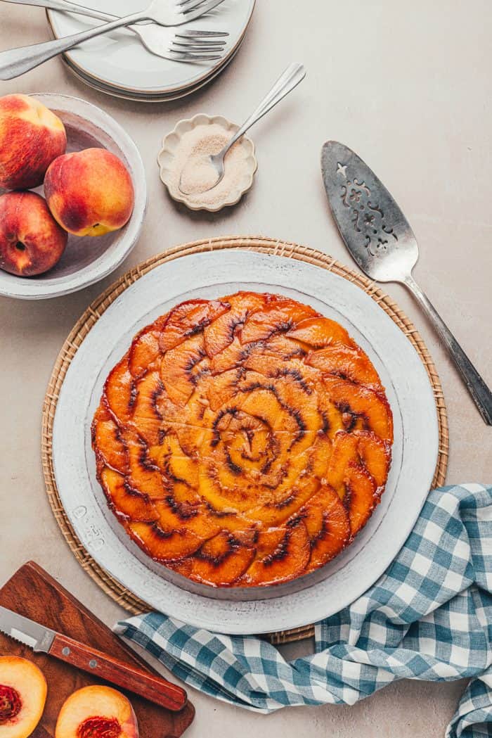 An upside-down peach cake on a white plate with a blue checkered towel and peaches scattered around.