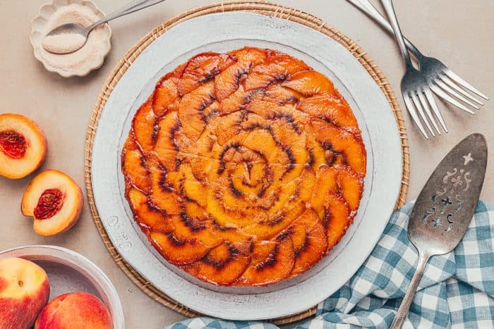 An upside-down peach cake on a white plate with a blue checkered towel and peaches scattered around.
