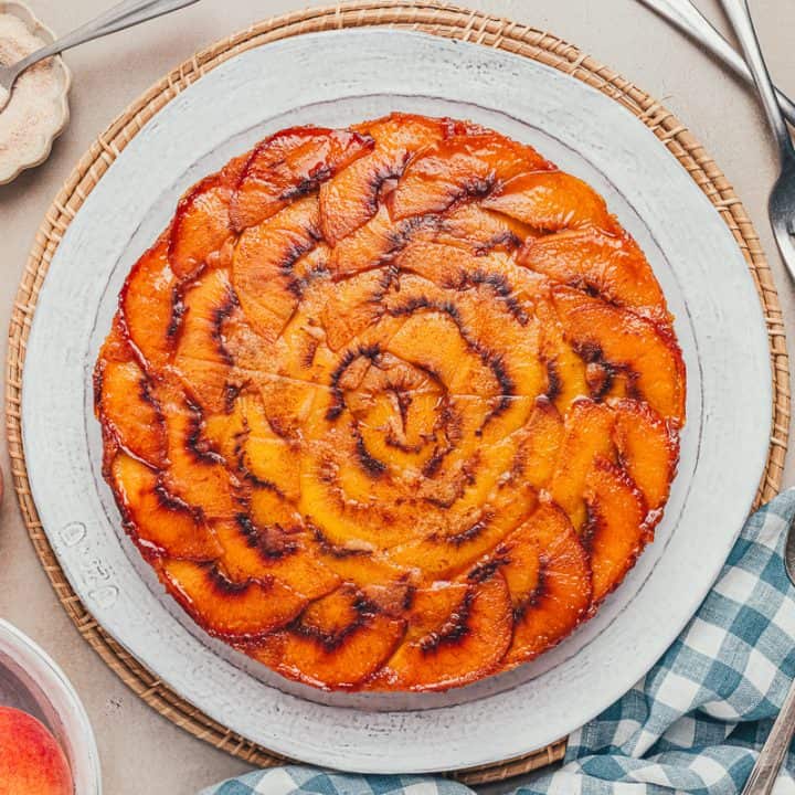 An upside-down peach cake on a white plate with a blue checkered towel and peaches scattered around.