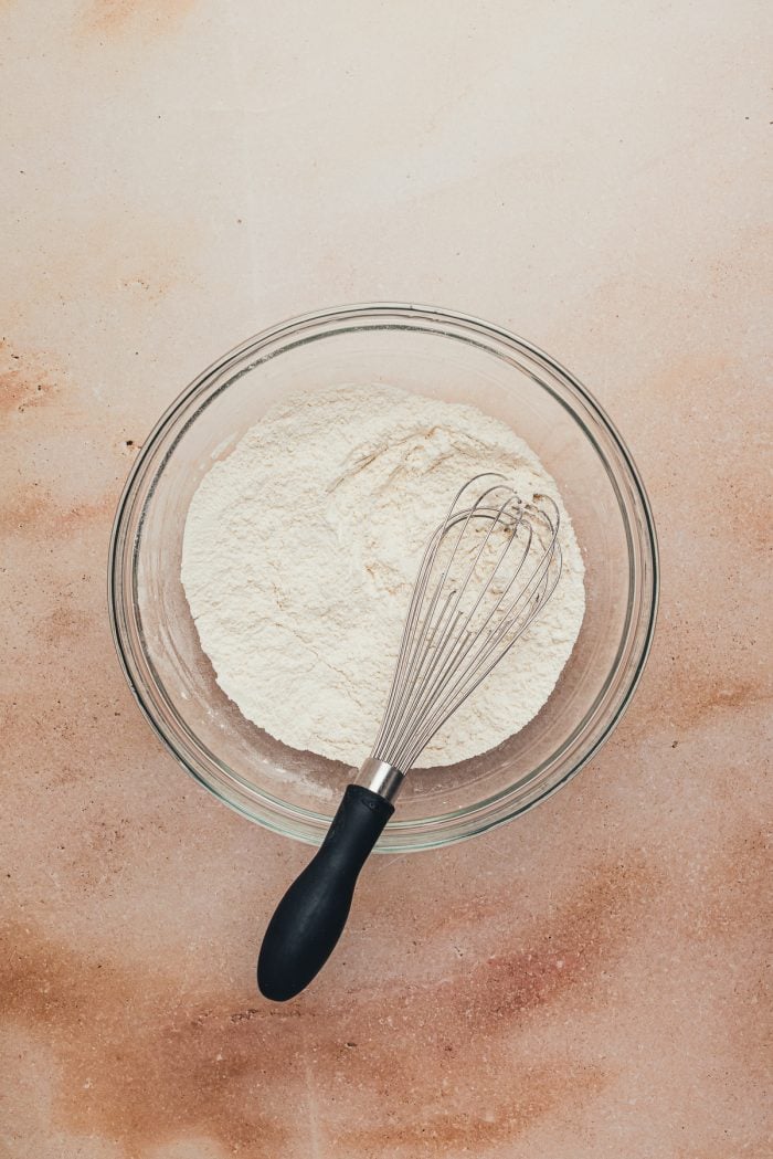 A glass bowl with flour in it and a whisk.