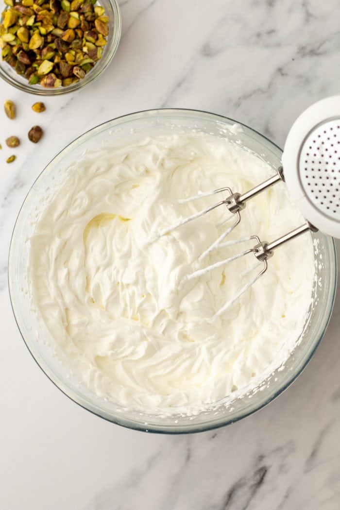A glass mixing bowl with whipped cream and a hand mixer.