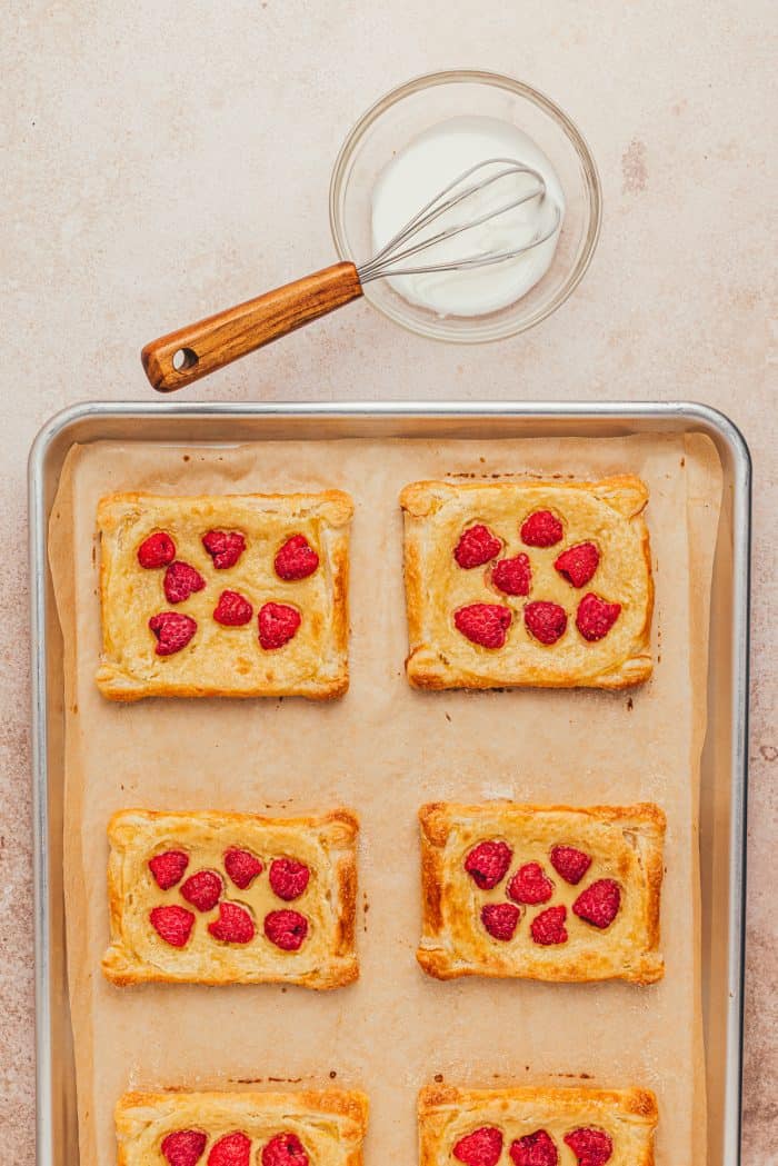 The danishes cooling on a baking sheet while a whisk is laying in a bowl of icing.