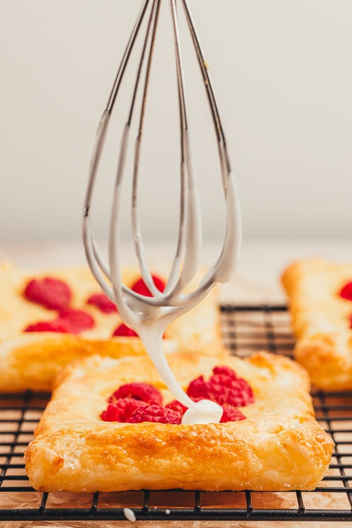 A whisk dripping icing over a freshly baked raspberry lemon cream cheese danish.