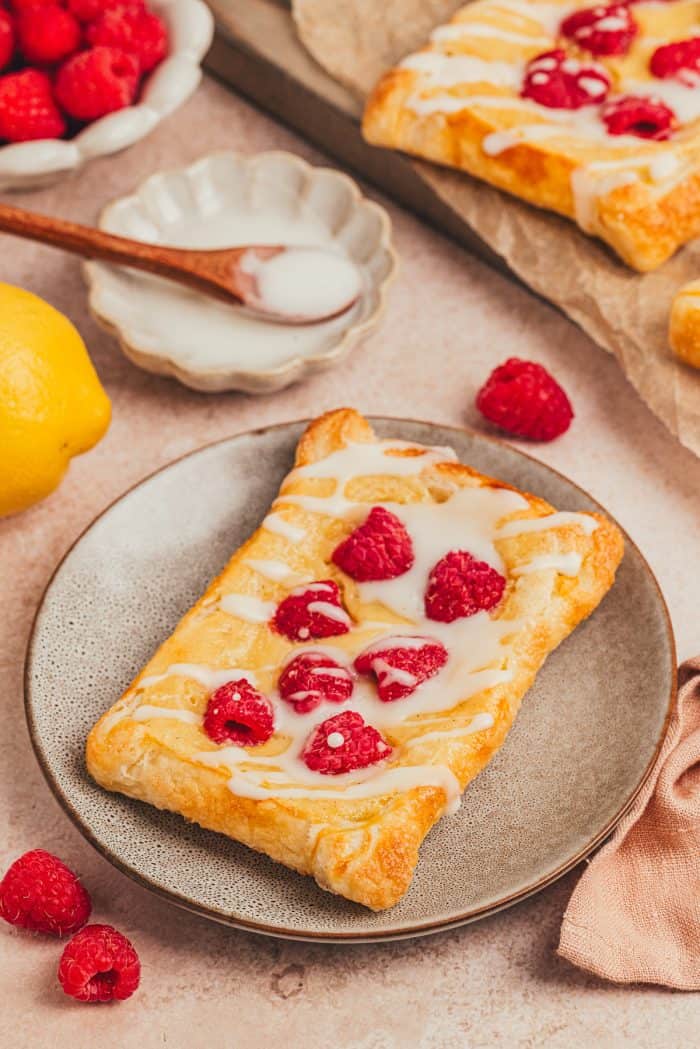 A homemade raspberry lemon cream cheese danish on a plate with a small bowl of icing with a spoon. 