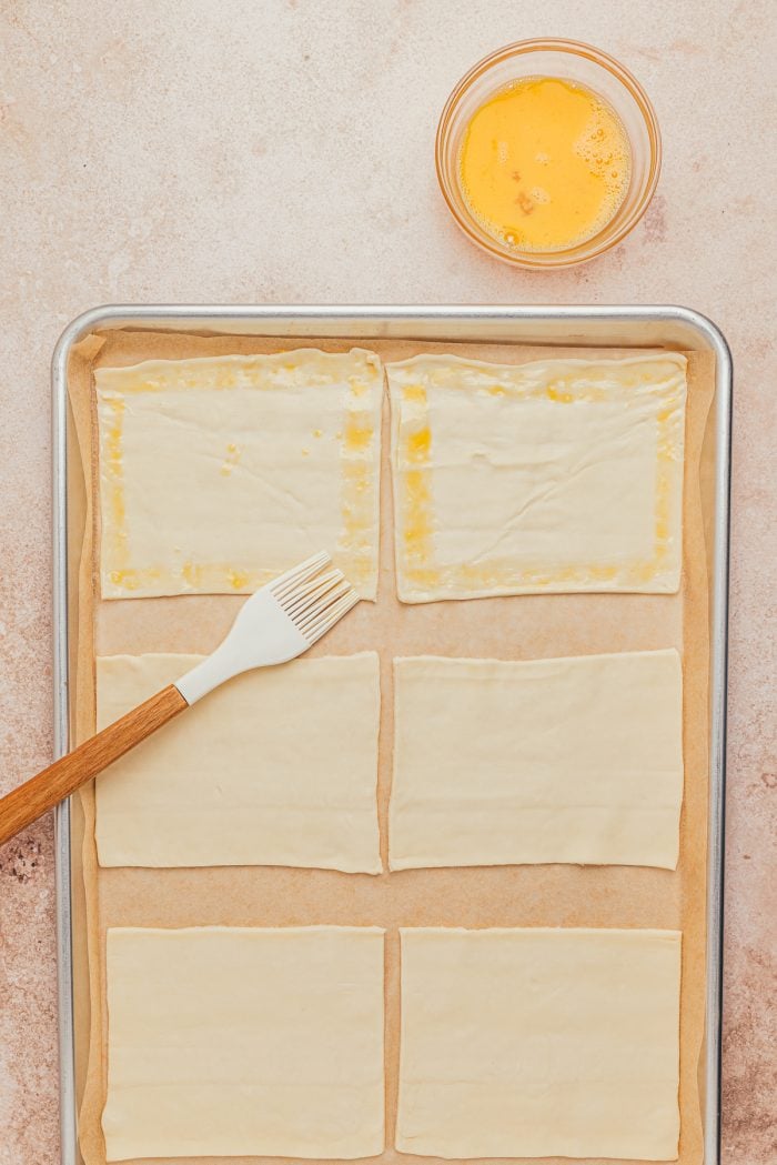 Egg being brushed over the edges of raw puff pastry on a baking sheet.