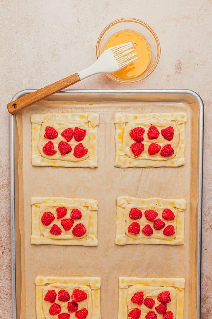 A baking sheet with unbaked cream cheese danishes and a pastry brush with a bowl of egg mixture.