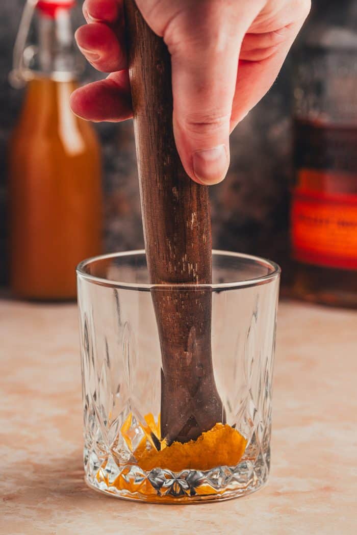 A muddling stick muddling the orange peel, bitters, and syrup.