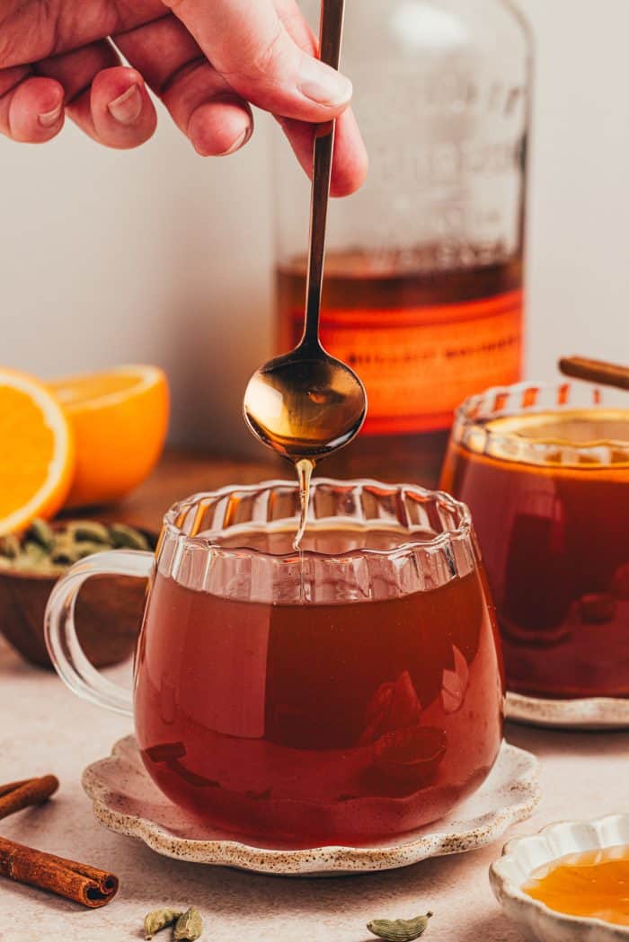 A glass mug filled with chai vanilla bean hot toddy and a spoon being dipped into it.
