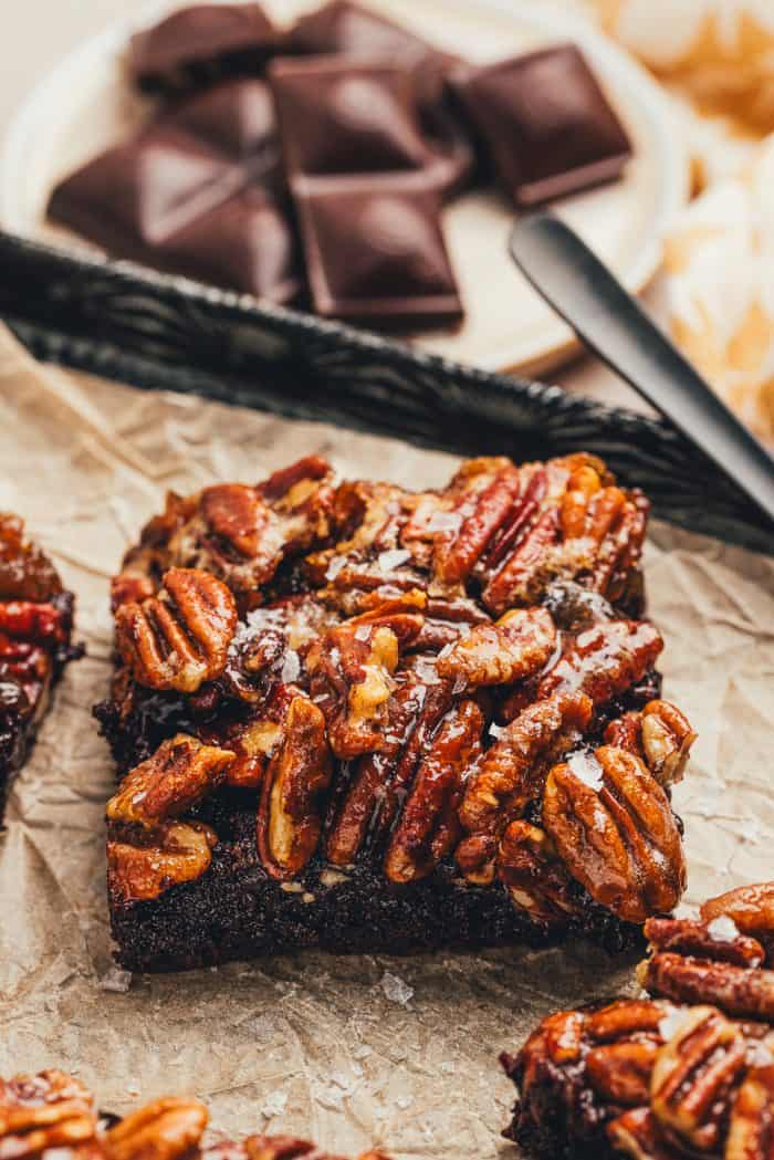 A single pecan pie brownie is on parchment paper with chocolate in the background.