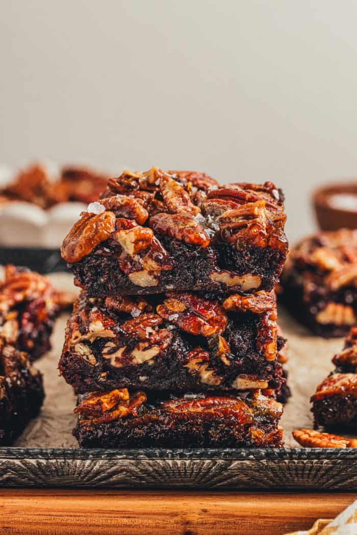 A stack of 3 brownies with pecan pie topping with various brownies in the background. 