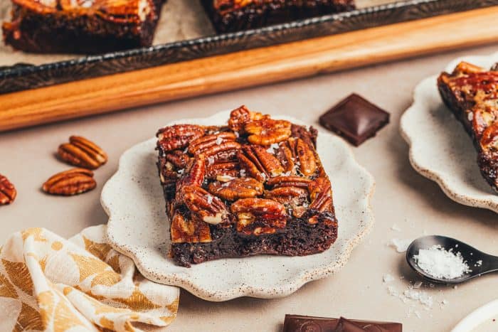 A brownie with a pecan pie topping is on a white plate with pecans scattered around it. 