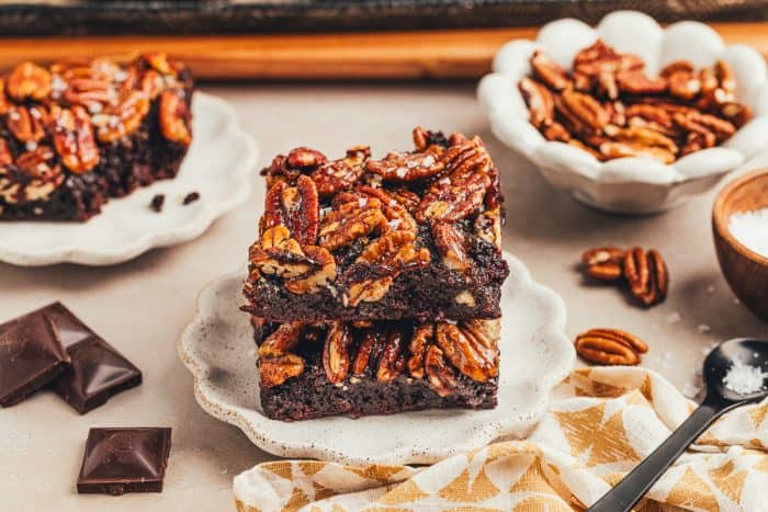A brownie with a pecan pie topping is on a white plate with pecans scattered around it. 