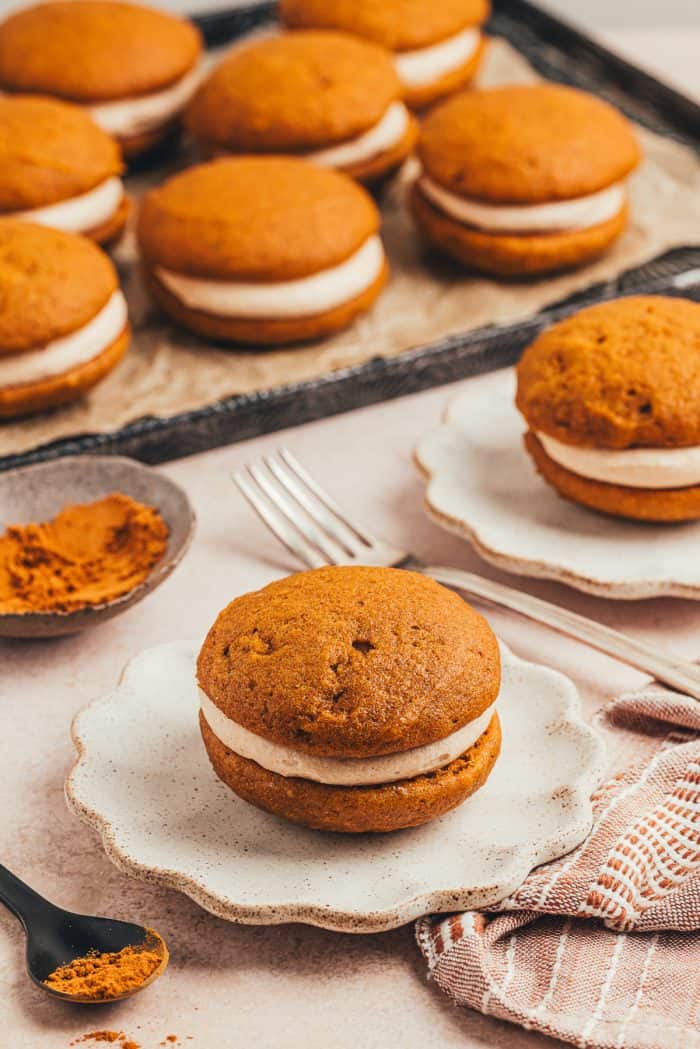 A pumpkin whoopie pies on a ruffled white plate with baking tray of pumpkin whoopie pies. 