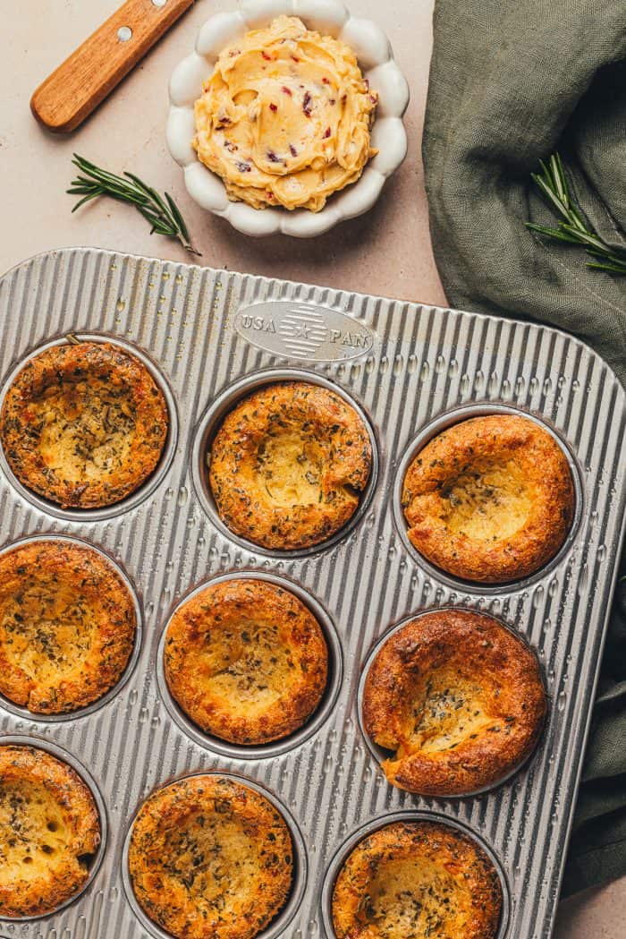 Herbed popovers in a baking tray with a green napkin and a bowl of butter in the background.