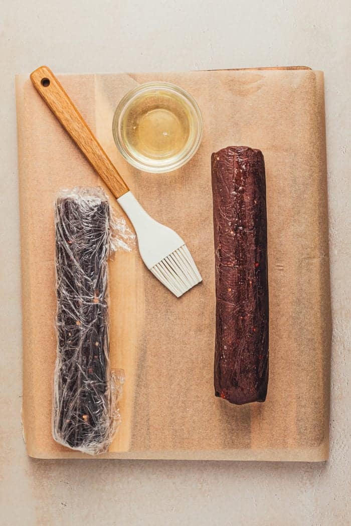 One log of dough wrapped in Saran wrap, with a brush and the other log of dough unwrapped.