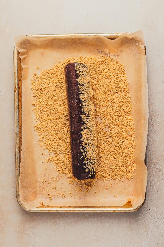 A log of cookie dough being rolled in finely chopped hazelnuts.