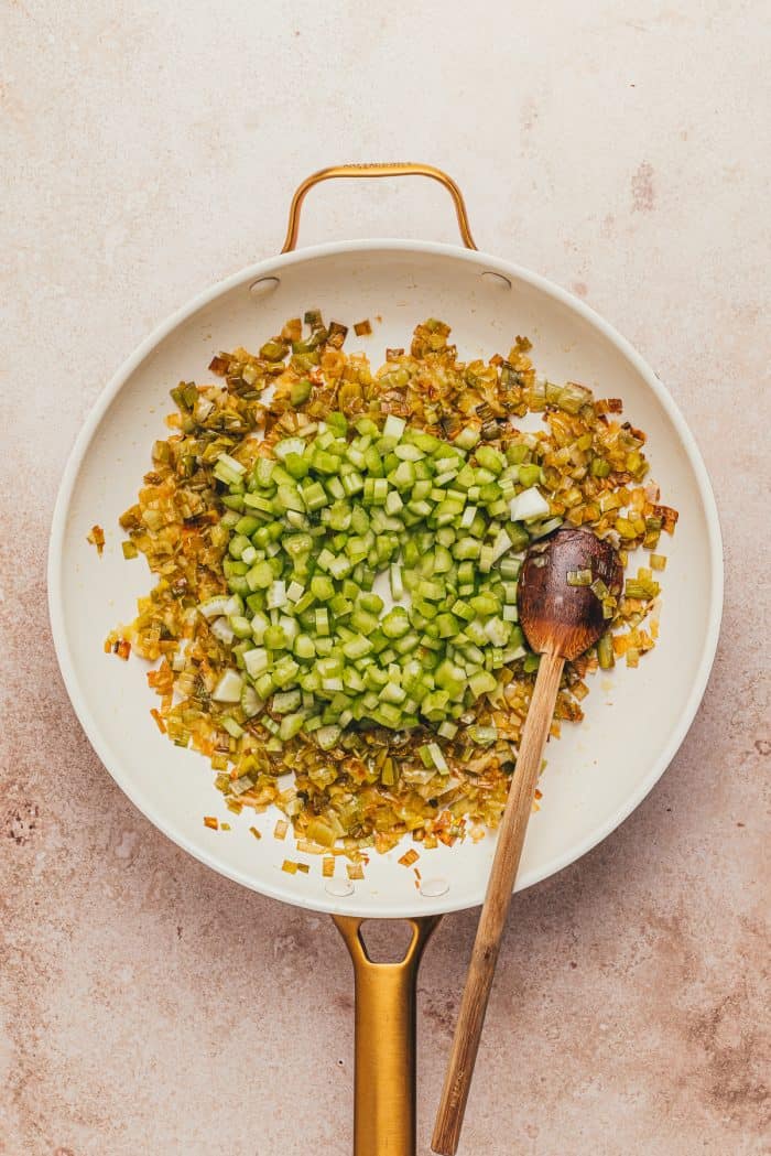 Caramelized leeks and diced celery in a white skillet with a wooden spoon.