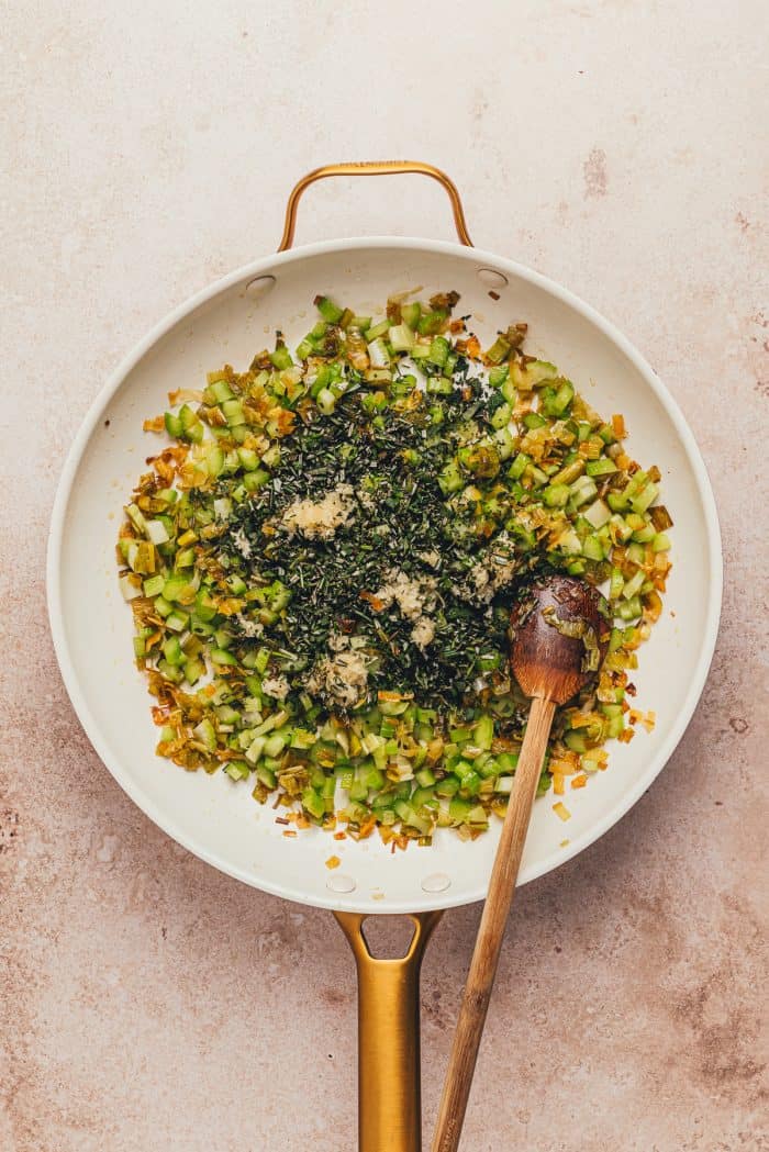 A white skillet with the leeks, celery, garlic, and herbs and a wooden spoon.