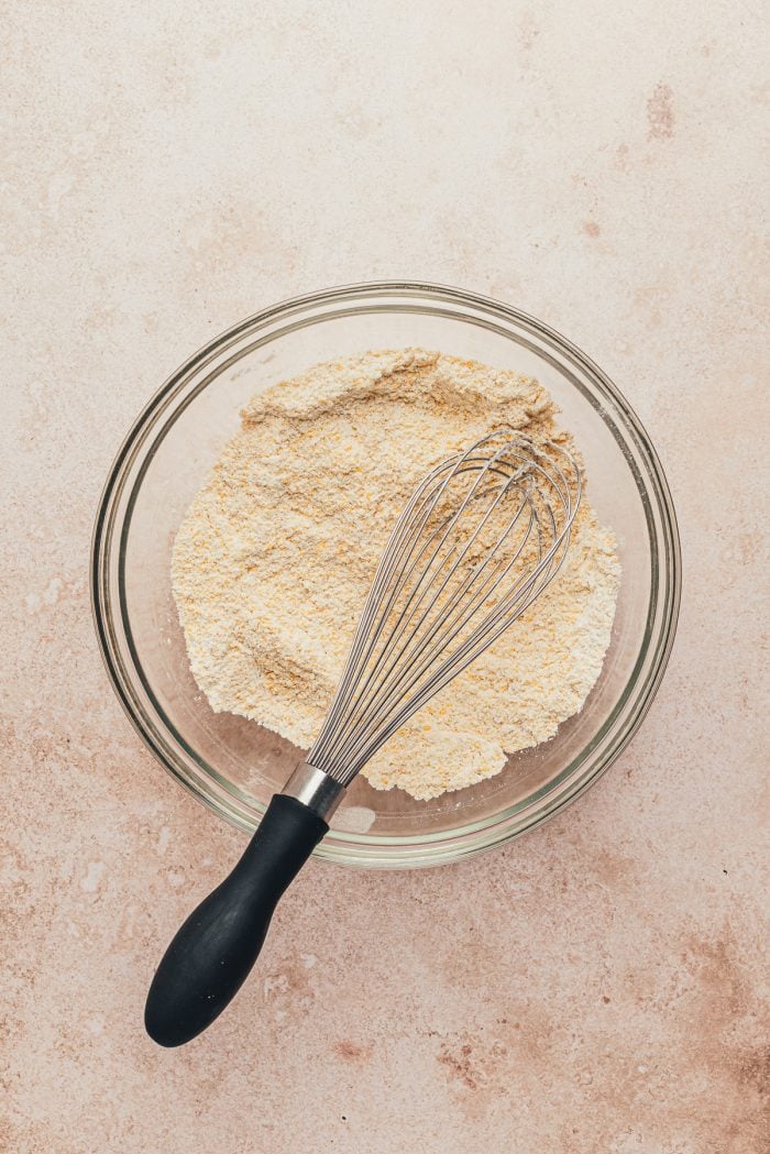 A glass bowl with cornmeal and flour and a whisk.