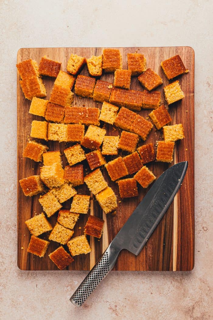 Cornbread cut into cubes on a wooden cutting board with a large knife.