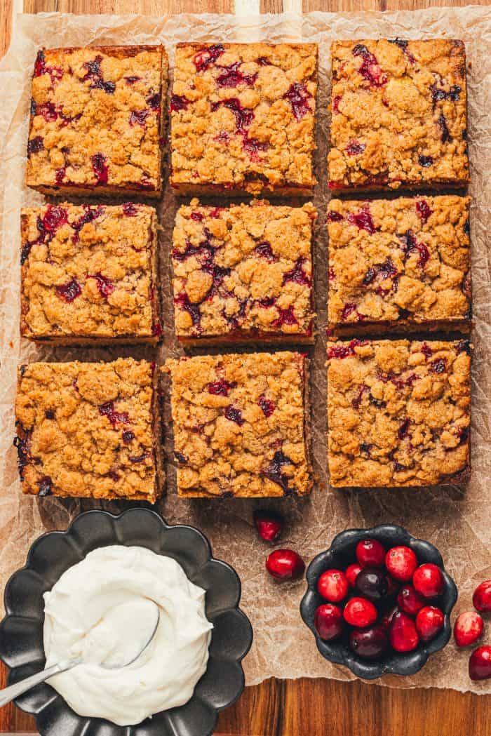 The top view of a cranberry crumb cake sliced with a bowl of creme fraiche and a smaller bowl of cranberries.