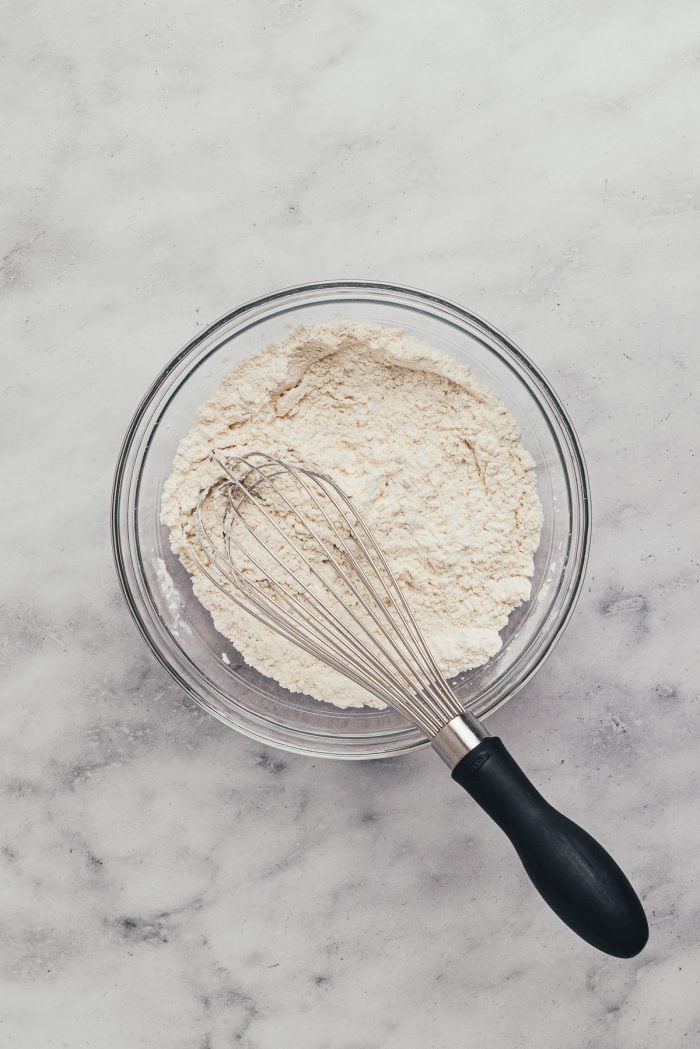 A glass mixing bowl with dry ingredients like flour and a whisk,