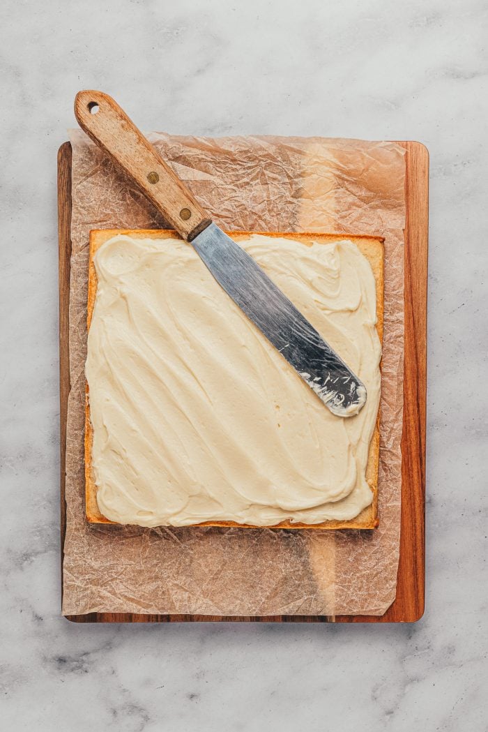 Baked sugar cookie bars on a wooden cutting board with the frosting being spread over the top.