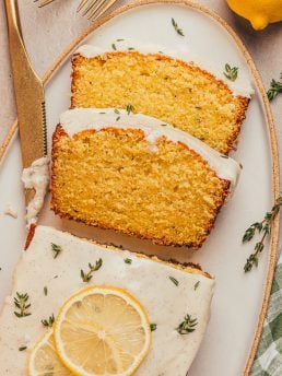 A platter with a loaf of lemon pound cake with slices of the cake and fresh thyme sprinkled around it.