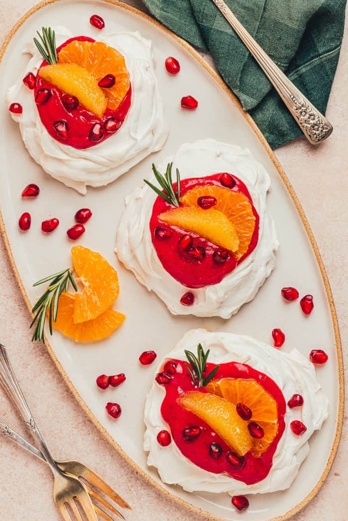 Three mini pavlovas with cranberry curd and orange slices with other garnishes on a platter.