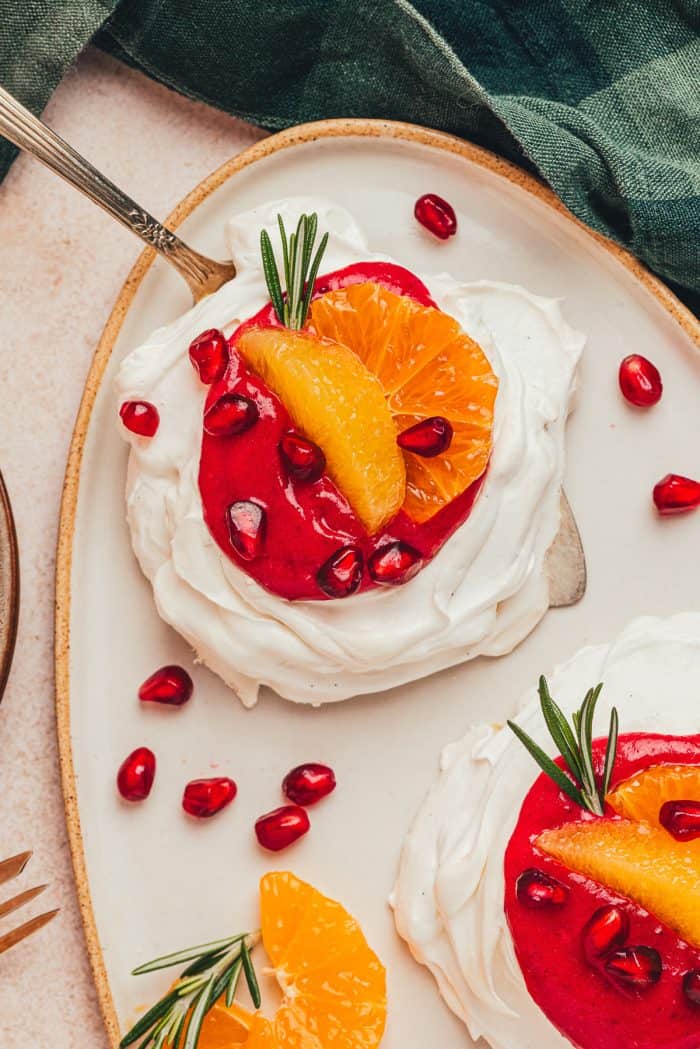 Two mini pavlovas with cranberry curd and orange slices with other garnishes on a platter with a cake spatula under one pavlova lifting it.