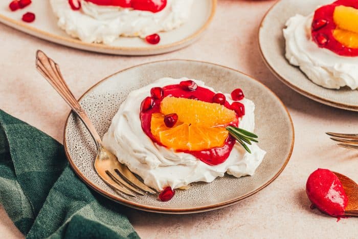 A mini pavlova with cranberry curd and orange slices with other garnishes on a plate with a fork.