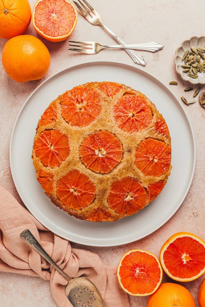 An Orange Cardamom Cake on a platter with oranges in the background.