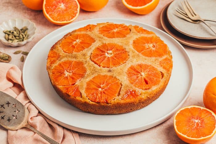 An Orange Cardamom Cake on a platter with oranges in the background.