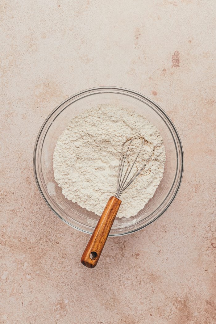 A glass bowl with flour and other dry ingredients and a whisk.
