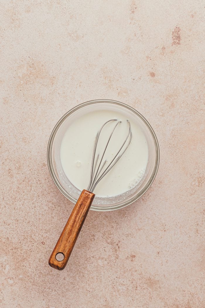 A glass bowl with sour cream and milk and a whisk.