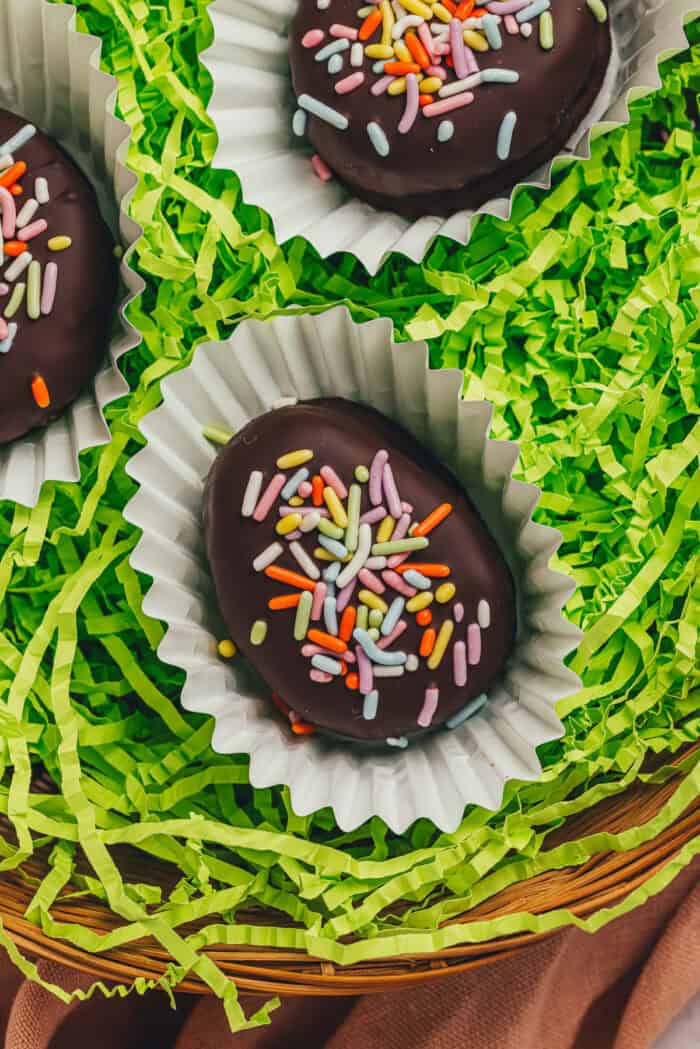 A few homemade chocolate-covered Easter eggs in confetti papers in a wicker basket with shredded green filling.