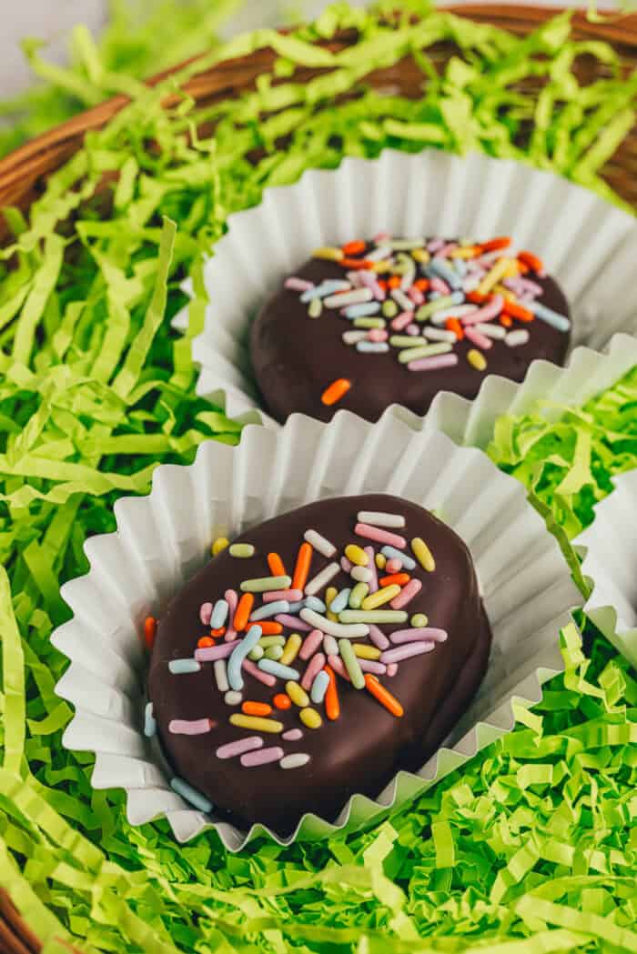 A few homemade chocolate-covered Easter eggs in confetti papers in a wicker basket with shredded green filling.